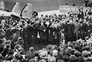A large crowd on an airfield; British Prime Minister Neville Chamberlain presents an assurance from German Chancellor Adolf Hitler.