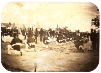 An old photograph showing a procession passing between lines of soldiers with tents in the background