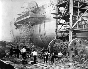 An old photograph showing a large iron paddlewheel ship being launched sideways, with workmen thrusting large baulks of timber under a large drum of iron chains