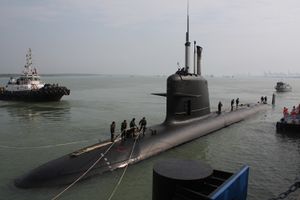 A Scorpène class submarine at dock, half out of the water. People on top are mooring it, and a boat can be seen in the background