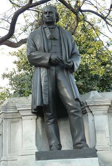 A bronze metal sculpture of a nineteenth century man wearing a long jacket or coat, trousers, waistcoat, with draughtsman's tools in his hands