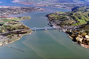 Rolling hills covered partially with buildings and roads frame the banks of a large channel of opaque water, which connects two larger bodies of water at the top left and bottom.