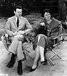 A black and white photograph of Alan J. Pakula seated next to Harper Lee in director's chairs watching the filming of To Kill a Mockingbird