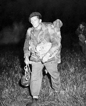 man in British Army uniform, carrying a parachute helmet and wearing a beret, other men can just be seen in the dark background