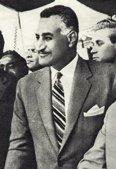 A smiling man with a black mustache faces the viewer's left. His hair is dark and short, white at the temples. He is wearing a western-style two-piece gray suit and white shirt, with an angularly striped tie and visible white pocket handkerchief. Behind him several faces are visible.