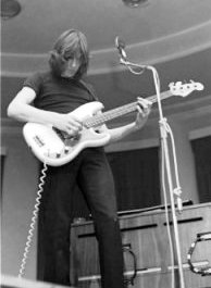 A monochrome image of Roger Waters playing bass guitar. He has shoulder-length hair, black attire, and is standing in front of a microphone.
