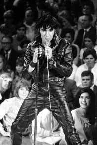 Presley, wearing a tight black leather jacket with upturned collar, black leather wristbands, and black leather pants, holds a microphone with a long cord. His hair, which looks black as well, falls across his forehead. In front of him is an empty microphone stand. Behind, beginning below stage level and rising up, audience members watch him. A young woman with long black hair in the front row gazes up ecstatically.