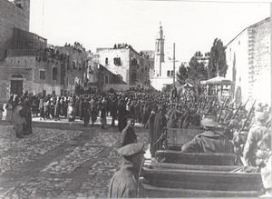 City buildings, some with people looking down from roofs, people and soldiers in a large square, with motor car in foreground