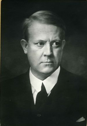 A black and white photographic portrait of a man aged around thirty, looking slightly to his left. He is dressed in a dark suit and tie; his hair is neatly combed into a parting.