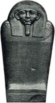 A dark stone coffin with a human face, the coffin stands upright facing the viewer.