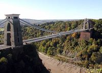 Suspension bridge between two brick built towers, over a wooded gorge, showing mud and water at the bottom. In the distance are hills.
