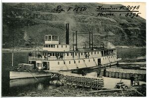 A steamboat waits at a pier on a river bank while sacks of cargo are loaded.