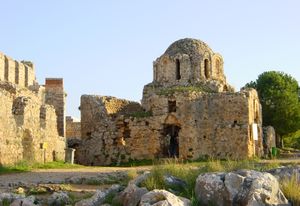 Ruins of a small stone domed structure built in a Byzantine style with tall windows. Grasses grow on the second level, as do trees behind it.