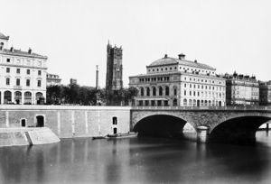 A city riverside scene with a bridge to the right. On the far bank are several buildings, the line of which is broken by a tall tower.