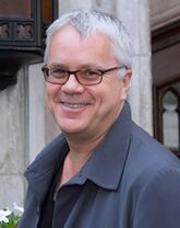 A standing caucasian man with short white and grey hair, wears glasses and a blue coat: He faces left towards the camera smiling.