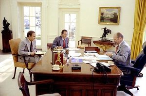 A man sits at his desk, smoking a pipe, while two other men speak to him from the other side of the desk.