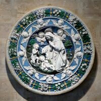 A circular terracotta plaque, sculptured in relief and glazed in intense colours of blue and green with white figures and motifs. At the centre the Virgin Mary, watched by John the Baptist, kneels in adoration of the baby Jesus. Little cherubs look on