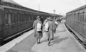 Soldiers walk down a station platform, while on either side of the platform are two trains. The men are wearing slouch hats and are carrying bags over their shoulders.