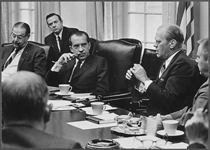 Men in suits are shown meeting in a conference room. Five men are shown, one of whom is speaking to man on his right. A sixth man is visible from behind, facing the speaker.
