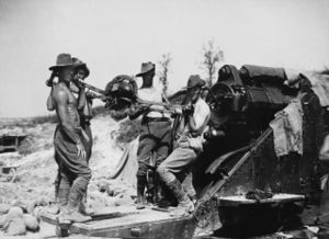 Bare-chested men in slouch hats and breeches stand by a large artillery piece, ready to load a shell.