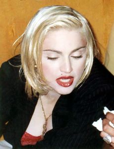 The image of a young blond woman. She is wearing a black coat. Her hair is short, straight and parted from the left to the right. She has bright red lips and appears to be speaking to someone on her left while looking down.