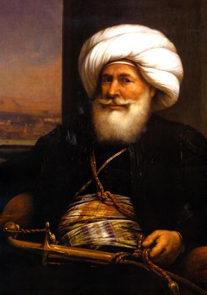 A man with a full white beard and long trained mustache faces the viewer. He wears a white turban and back robe. High on his waist is a gold sash decorated with purple and orange stripes. His left hand holds a cord that goes across his chest, and is connected to a sheathed sword in front of him.