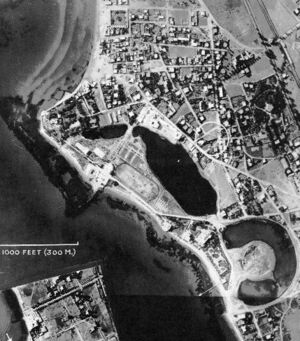 A black and white aerial photograph of an urban area by the sea with a water-filled, torus-shaped inlet.