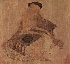 Elderly Chinese man, seated at a low writing desk and holding a scroll
