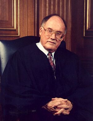 Seated portrait, from waist up, of a man in black robes, coat, and tie. He wears glasses and has a receding hairline. His hands are folded.