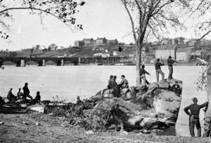 Black-and-white photo of several military men idling on a river bank. Across the river are several large buildings.