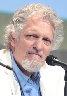 A white man with a short white beard, and white and grey curly hair: He wears a white suit with a blue shirt. The man stares away from the camera looking angry.