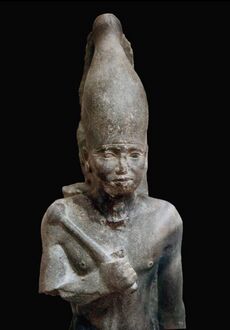 Statue of a standing pharaoh in black stone. He is holding a mace and a rounded crown.