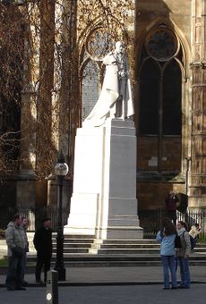 White stone statue of George V in Garter robes on a plinth of the same stone standing in a street