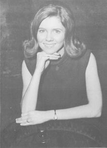 Black and white photograph of a young woman with 1960s style hair poses for the camera with a smile and her right hand under her chin