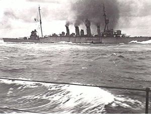 A large four-stacked warship billowing thick black smoke and moving through moderate seas