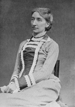 A photograph of a dark-haired white woman of about 40 sitting in an upright chair. She wears a long sleeved, collared dress with decorative material down the front and at the cuffs and neck.