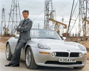 Bond in a grey suit, leaning against a roadster with oil rigs in the background.