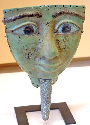 An oversized, shallow mask depicting a large face. The face is roughly oval-shaped, but the top of the mask is a horizontal line just above the eyebrows, leaving the entire mask roughly triangular. The entire face is flattened, but the bulbous nose protrudes away from the face. The eyes are large and almond shaped, and both the eyes and braided eyebrows are disproportionally large in comparison with the mouth, which has full lips. The front of the face is clean-shaven, but below the chin, there is a long, narrow, pointed, braided false beard that was characteristic of ancient Egyptian royalty.