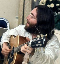 A bearded, bespectacled man in his late twenties, with long black hair and wearing a loose-fitting white shirt, sings and plays an acoustic guitar. White flowers are visible behind and to the right of him.
