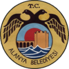 A dark-yellow circular seal with a smaller circle inside it that portrays a fortified tower and wall behind blue waves. The smaller circle is enclosed by a black two-headed bird with the text T.C. above and Alanya Belediyesi below.