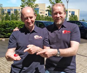 Tim (right) and Chris (left) Stamper; the middle-aged men both wear black polos with red logos on the right of the buttons. Tim has a beard, while Chris is balding.