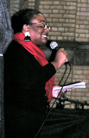 50ish woman in black, with a bright red scarf, large black-and-white earrings and glasses, smiling and holding a microphone in one hand and a sheet of paper in the other