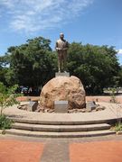 A bronze-colored, life-size statue of a prominent black man in a suit, his hands in front of his stomach. The statue is on top of a pedestal which is on top of a large brown rock, surrounded by smaller rocks. The statue has trees behind it, and it is in a small brick-paved square.