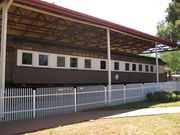 A brown train, viewed from the side, is at rest under a small open station. “1035 Rhodesia Railways” is written in gold letters over the train's windows. There is a white fence and a brick walkway in front of the train.
