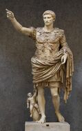 A marble statue of the Emperor Augustus. He stands with one arm raised as if in command. Augustus is portrayed as a man of about thirty five, with short hair and clean shaven. He wears Roman military uniform of a breast plate, leather accoutrements and a cloak over a short tunic. The breastplate is decorated with symbolic figures. As a work of art, the statue displays high technical mastery.