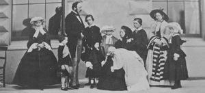 Photograph of a seated Victoria, dressed in black, holding an infant with her children and Prince Albert standing around her