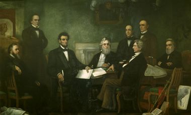 A dark-haired, bearded, middle-aged man holding documents is seated among seven other men.]]