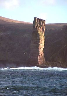 A tall perpendicular stack of brown rock stands in the sunlight in front of a shore with high cliffs that lie in the shadows.