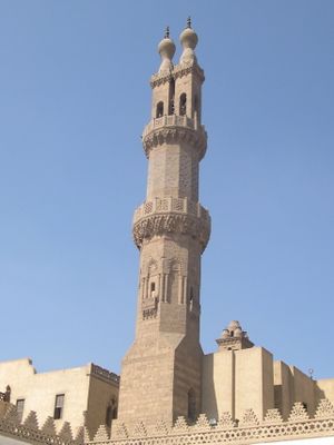 An ornate carved stone octagonal minaret, with a carved stone railing around balconies at its center and near its top. Above the second balcony the minaret splits into two rectangular shafts, each tipped by railing and a bulb-shaped finial.