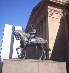 Equestrian statue in dark grey metal of George V in military dress uniform on a plinth of red granite outside a Classical building of red sandstone